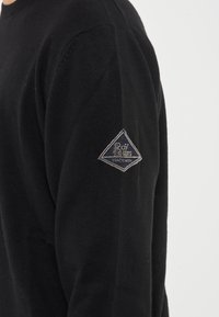 Maglione nero a maglia con scollo rotondo e maniche lunghe. Presenta una toppa ricamata a forma di diamante sul braccio sinistro.