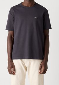 Czarny t-shirt z krótkim rękawem z bawełny w kolorze ciemnoszarym, okrągły dekolt, z małym białym logo po lewej stronie klatki piersiowej. Noszony z jasnymi spodniami.