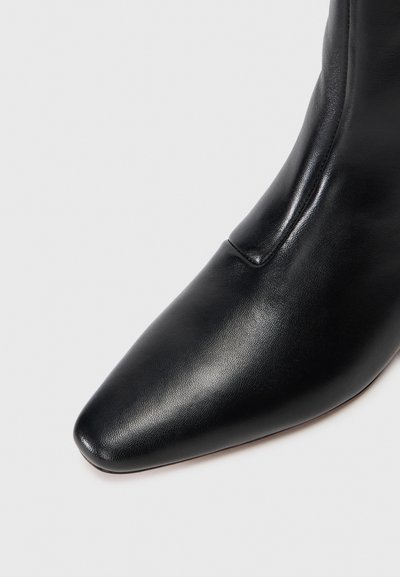 Stivale alla caviglia in pelle nera con punta affusolata, texture liscia, cuciture dettagliate e un pannello elastico laterale su uno sfondo semplice.