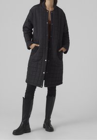 Mujer con un abrigo acolchado negro de longitud hasta la rodilla con botones a presión, pantalones skinny negros y botas negras hasta la rodilla, con las manos en los bolsillos.