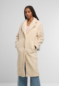 Cremfarbener Fleece-Mantel mit Reverskragen, seitlichen Taschen und einer weichen, strukturierten Oberfläche. Besitzt eine knöpfellose Vorderseite und eine entspannte Passform.