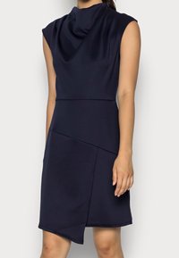 Ärmlös klänning i marinblått med draperad halsringning och asymmetrisk nederkant, buren av en stående person mot en neutral bakgrund.
