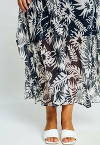 Kvinnas underben och hand som bär en genomskinlig marinblå och vit blommig kjol samt vita öppna sandaler med häl på en vit bakgrund.