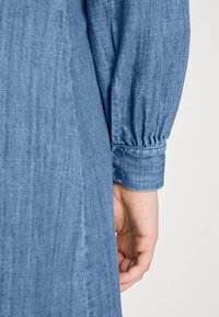 Sukienka dżinsowa z rękawami, charakteryzującymi się miękkim, teksturowanym materiałem, bufkowatym fasonem oraz dwoma guzikami na mankiecie w średnim odcieniu niebieskiego.