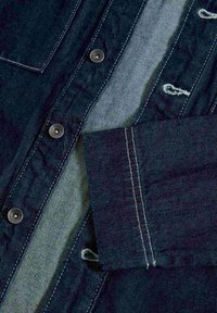 Camisa de mezclilla de tejido azul oscuro con subrayado en azul claro contrastante. Detalles de botones notables y acentos de costura visibles.