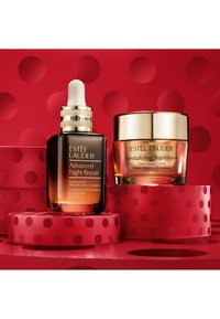 Estee Lauder Hautpflegeprodukte: dunkelglasiger Tropferflasche und Creme-Glas mit goldenen Deckeln, vor einem hellroten Hintergrund mit Punkten.
