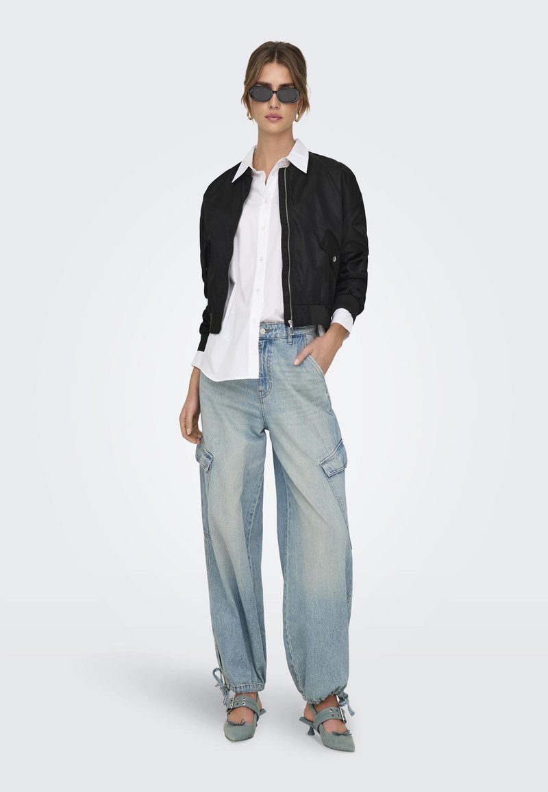 Black bomber jacket over a white button-up shirt, paired with light blue cargo jeans and gray heeled mules. Accessories include dark sunglasses.