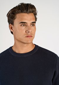 Marineblaue Rippstrickpullover mit Rundhalsausschnitt, ausgestattet mit einem strukturierten Muster und figurnaher Passform. Der Stoff wirkt weich und elastisch.