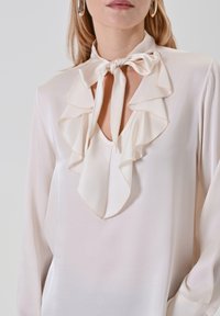 Blusa di seta bianca sporca con scollatura a V profonda, caratterizzata da un colletto arricciato e un fiocco annodato al collo. Le maniche lunghe completano il design.