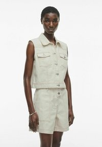 Chaleco de denim beige claro con diseño sin mangas, botones de presión en la parte delantera y dos bolsillos en el pecho. Combinado con pantalones cortos de cintura alta a juego.