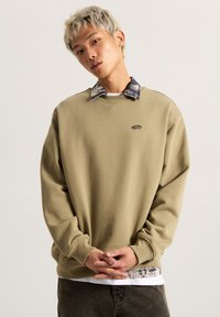Suéter caqui con cuello redondo y corte relajado, que presenta un logo negro y un cuello en capas con un patrón sutil. Tejido texturizado, puños acanalados.