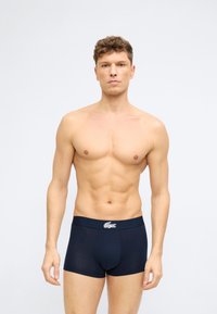 Νεαρός άνδρας χωρίς μπλούζα, με κοντά σγουρά μαλλιά, φορώντας σκούρα μπλε εσώρουχα boxer με λευκό λογότυπο στη μέση, στέκεται μπροστά σε απλό φόντο.