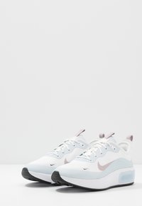 Valkoiset urheilukengät, joissa on vaaleansiniset yksityiskohdat, teksturoitu pohja ja vaaleanpunainen kantalenkki; sisältää swoosh-logon ja nauhakiinnityksen.