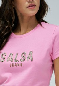 Camiseta rosa de manga corta con cuello redondo que presenta el texto "SALSA" y "JEANS" adornado con cristales en la parte delantera. Tejido de algodón suave.