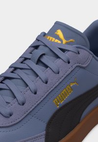 Κοντινή λήψη μπλε αθλητικού παπουτσιού Puma με μπλε κορδόνια, μαύρη λωρίδα στο πλάι και χρυσό λογότυπο Puma στη γλώσσα και στο πλάι.