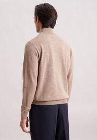 Mann in beigem Rollkragenpullover und marineblauen Hosen, der mit dem Rücken zu einer schlicht hellen Hintergrund steht.