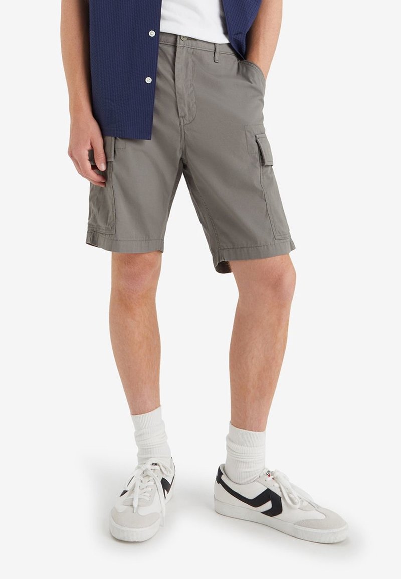 Shorts cargo gris avec poches latérales, en tissu léger, présentant une coupe décontractée et des ourlets retroussés, portés avec des baskets blanches et des chaussettes.