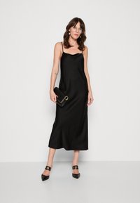 Robe nuisette en satin noir avec de fines bretelles, présentant une texture lisse et une silhouette fluide. Associée à une pochette noire et des escarpins pointus.