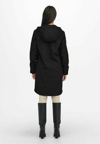 Una mujer está de pie con un largo abrigo negro con capucha, combinado con mallas beige y botas negras hasta la rodilla, de espaldas al espectador.