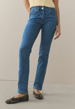 Nő középderékig érő, kék, egyenes szárú farmert és kígyóbőr mintás lapos cipőt visel, egy sima, világos háttér előtt áll.