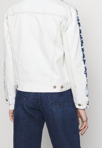 Bílá denimová bunda s indigo tie-dye akcenty na rukávech, standardním límci a kovovými knoflíky, spárovaná s tmavými denimovými džíny.
