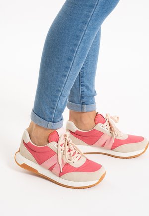 Person trägt hochgekrempelte blaue Jeans und rosa-beige Freizeitsneaker mit weißen Schnürsenkeln, steht vor weißem Hintergrund.