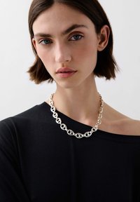 Stříbrný řetízek s náhrdelníkem, který se skládá z propletených oválných a kulatých oček. Modelka má na sobě černý top bez ramínek. Hladký, reflexní povrch.