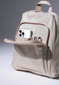 Zaino beige in tessuto texturizzato, con cerniera e tasca anteriore che contiene uno smartphone e delle penne. Manico morbido sulla parte superiore.