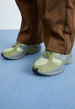 Green sneakers with mesh and suede panels, featuring a thick gray rubber sole and tonal laces. Worn with brown pants, on a blue surface.