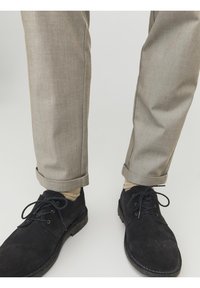 Béžové kalhoty na míru s ohrnutými nohavicemi, v kombinaci s černými semišovými šněrovacími botami. Oblečení má neutrální barevnou paletu a jednoduchý design.