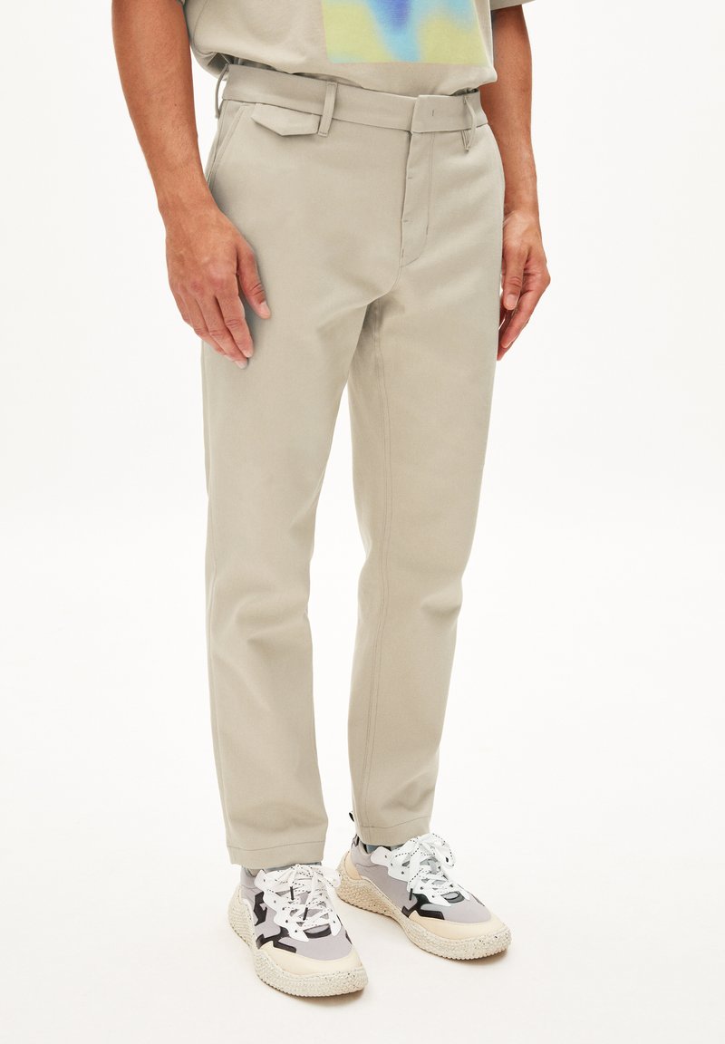 Beige Hosen im geraden Schnitt mit flachem Frontbereich und seitlichen Taschen, kombiniert mit gemusterten Sneakers mit schwarzen Akzenten und weißen Schnürsenkeln.