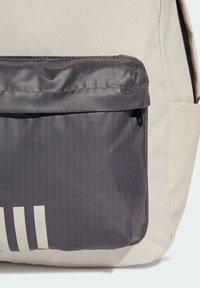 Zaino con un corpo in tessuto beige e una tasca frontale grigia texturizzata. Presenta tre strisce orizzontali bianche nella parte inferiore. Tasca con cerniera.