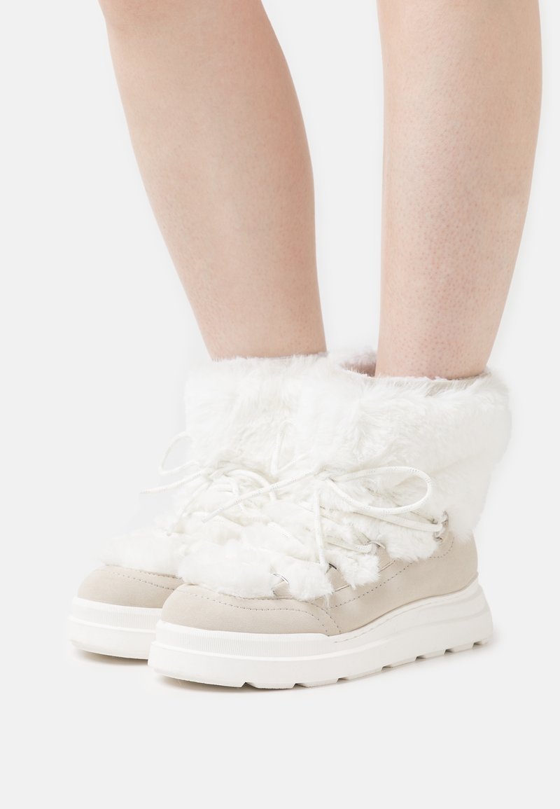 Bottes blanches en fourrure jusque la cheville avec des accents en daim beige, bout rond, lacets blancs et semelle blanche épaisse, avec un revers texturé et confortable.