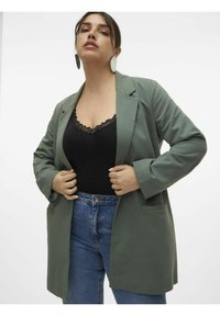 Blazer vert olive en tissu doux avec un design structuré, doté de deux poches avant et de manches retroussées, porté par-dessus un top noir avec des bordures en dentelle.
