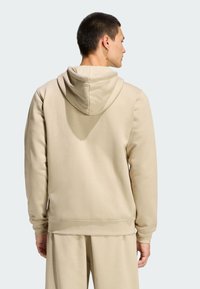 Béžová mikina s kapucňou so šnúrkami, hladkou textúrou a rebrovanými manžetami a lemom. Nosená s béžovými nohavicami rovnakej farby, ktoré majú voľný strih.