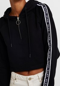 Černá zkrácená mikina na zip s kapucí a stahovací šňůrkou. Obsahuje bílé logo pruhy podél rukávů a kroužkový táhlo na zipu. Měkká textura.