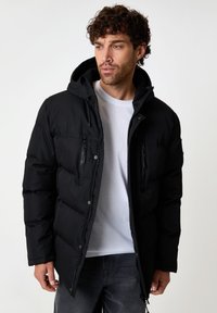 Veste en doudoune noire avec capuche, dotée de poches poitrine zippées et d'une fermeture éclair à l'avant. Tissu texturé avec une finition lisse et design isolé.