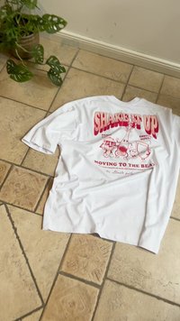White cotton t-shirt featuring a graphic print in red with playful characters and text reading "SHAKE IT UP" and "MOVING TO THE BEAT."