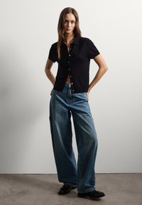 Mujer vestida con una camisa negra de botones de manga corta, jeans de pierna ancha azules y zapatos negros, de pie con las manos en las caderas frente a una pared lisa.