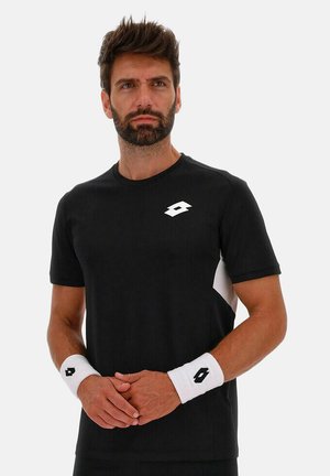 Mann trägt schwarzes Sport-T-Shirt und weiße Armbänder mit passenden Logos, steht vor hellem einfarbigem Hintergrund mit verschränkten Händen.