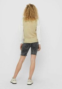 Gilet beige lavorato a maglia sopra una camicia bianca a maniche lunghe, abbinato a pantaloni corti in denim neri strappati. Calzature casual, sfondo chiaro.