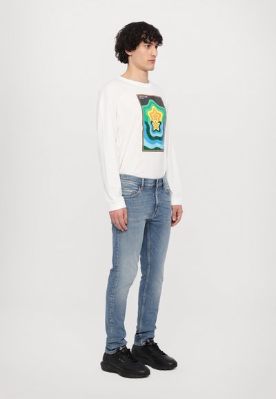 Wit overhemd met lange mouwen en een veelkleurig grafisch ontwerp, gecombineerd met lichtblauwe jeans en zwarte sneakers. Casual pasvorm en ontspannen stijl.
