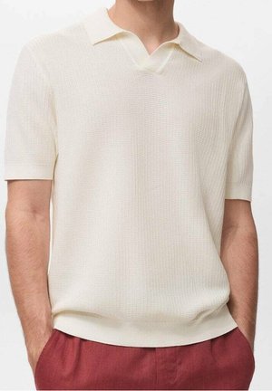 Homme portant un polo en maille à manches courtes de couleur blanc cassé, associé à un pantalon taille haute bordeaux, les mains dans les poches, debout devant un fond uni.