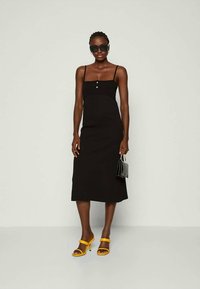 Mustreeritud must kleit õhukeste rihmadega, ruudukujulise kaelusega ja nööpidetailidega, koos kollaste kontsakingade ja musta käekotiga.