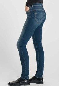 Jean skinny en denim bleu foncé, taille haute, avec poches arrière ornées d'un motif de couture décoratif et jambes ajustées.