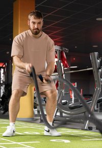 Athlète masculin en tenue de sport rose clair utilisant des cordes lourdes sur un sol de gymnase vert. Baskets blanches, équipement de gym en arrière-plan.