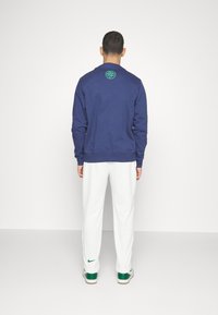 Mörkblå sweatshirt med långa ärmar och ribbade mudd, med en grön logotyp på baksidan. Matcher med vita sweatpants och gröna sneakers.