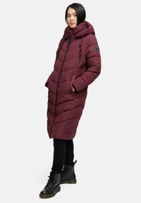 Lunga giacca puffa bordeaux con cappuccio, dotata di chiusura con zip, motivo a chevron e cordini. Abbinata a stivali neri e leggings.