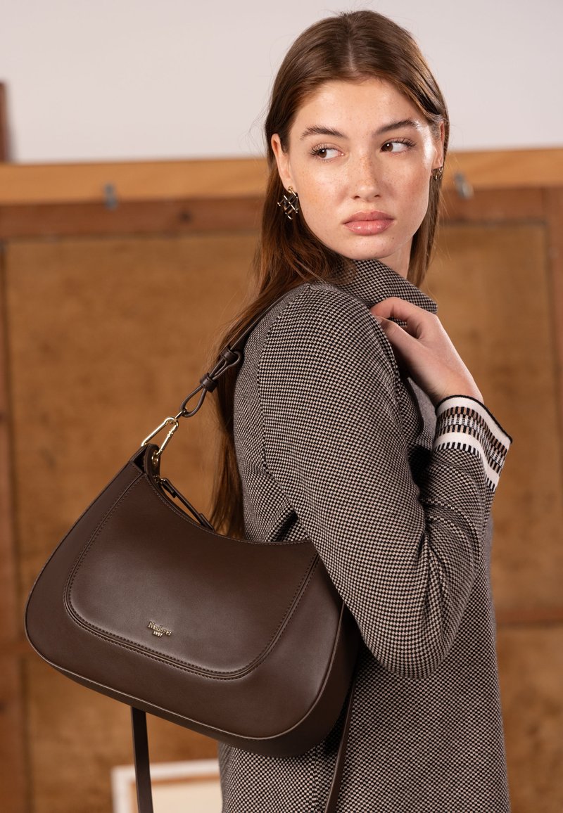 Bruine leren handtas met een gebogen vorm, voorzien van een goudkleurig metalen detail en een afneembare riem. Gestructureerde grijze kleding met patroon.