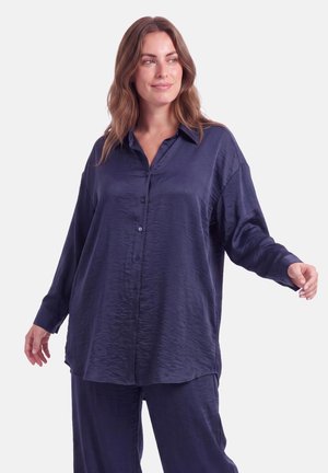 Vrouw met golvend haar die een losse, knoopsluitende marineblauwe satijnen blouse en bijpassende broek draagt, staand met licht uitgespreide armen.